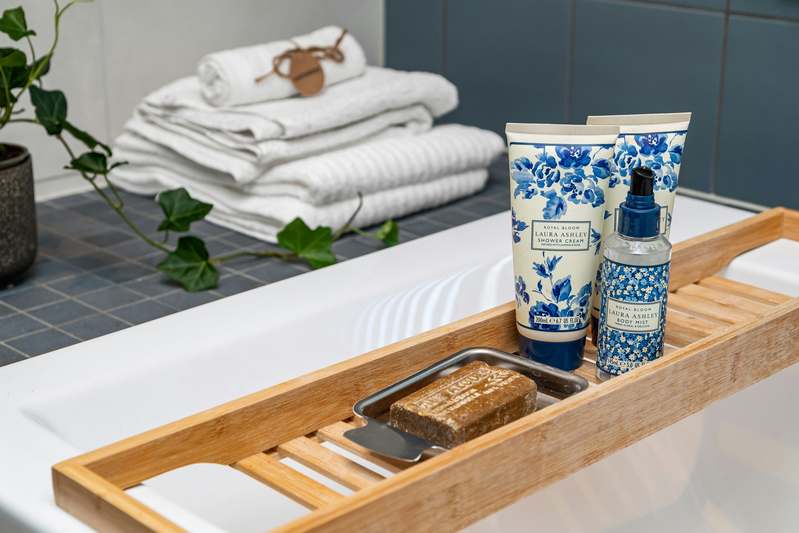 Close-up of hands pouring oat-infused bath milk into a steaming white bathtub, surrounded by eucalyptus leaves